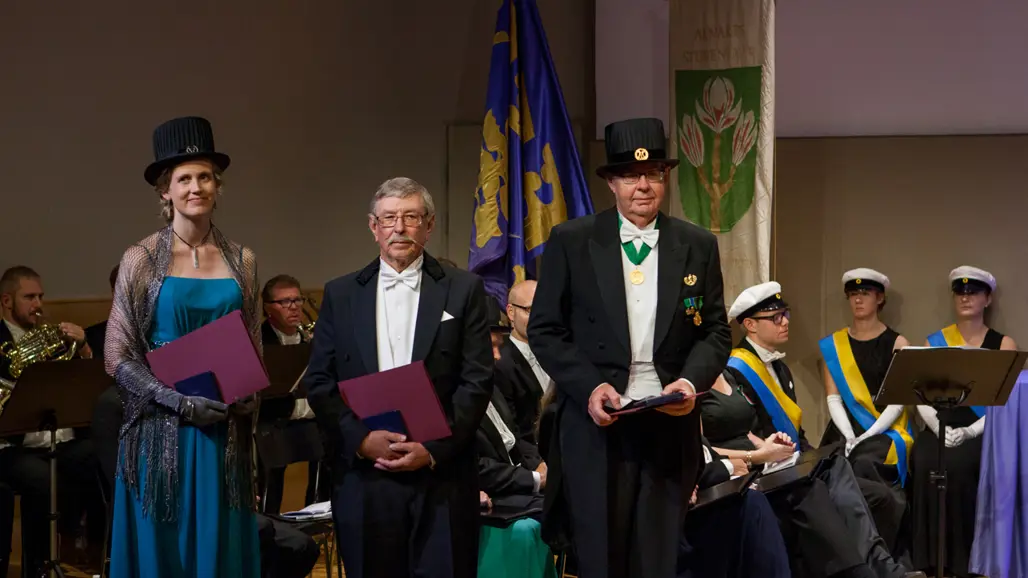 Tre personer i högtidskläder och doktorshattar står på en scen under en akademisk ceremoni, med musiker och fanor i bakgrunden.