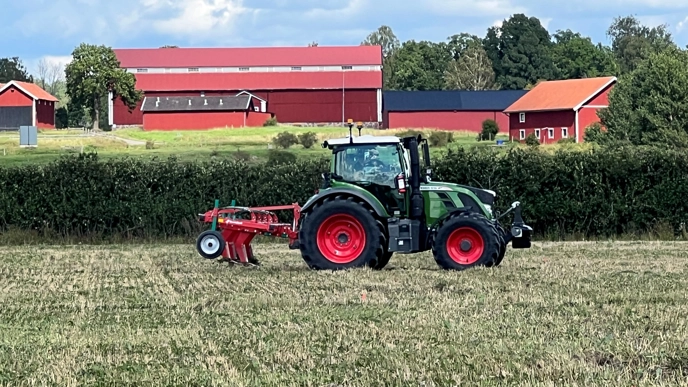 A tractor drives in a field. Red buildings are visible in the background.
