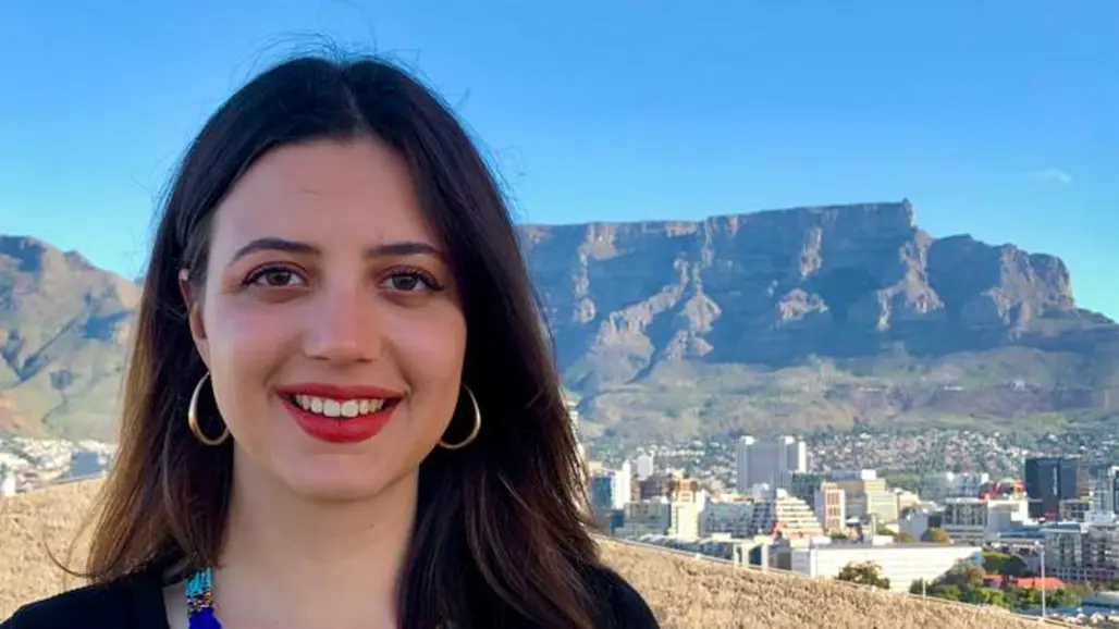 A smiling person with long dark hair, a colourful beaded necklace, and large earrings stands in front of a city with a distinctive flat-topped mountain in the background, on a clear day.
