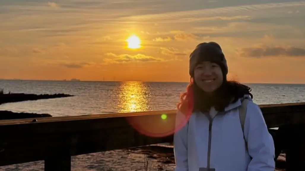 A person wearing a white jacket and beanie smiles at the camera by a pier, with the sea and a warm sunset behind them. The sun reflects on the water.