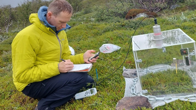 A man sits with a measuring instrument in a mountain environment. Photo.