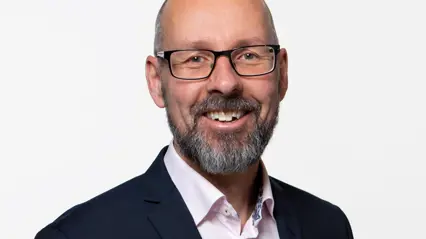 En person med kort, grått skägg och glasögon står mot en vit bakgrund. Personen bär en mörkblå kostym och en ljusrosa skjorta med ett mönstrat band runt halsen.