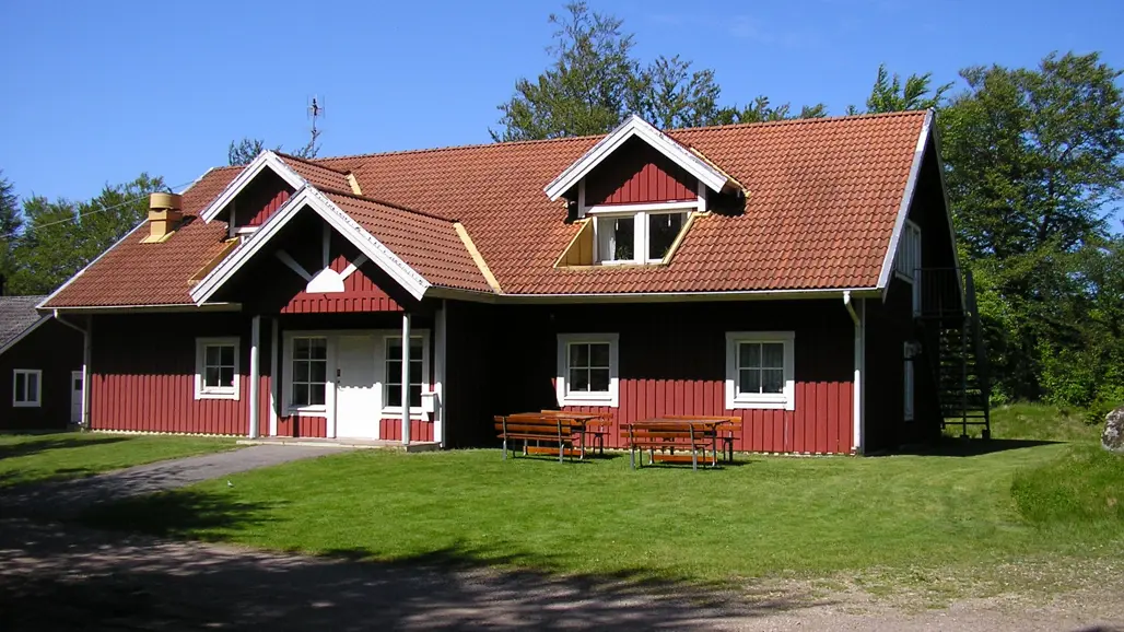 Ett rött tvåvåningshus av trä med tegeltak och vita knutar, på gräsmattan framför står två picknickbord.