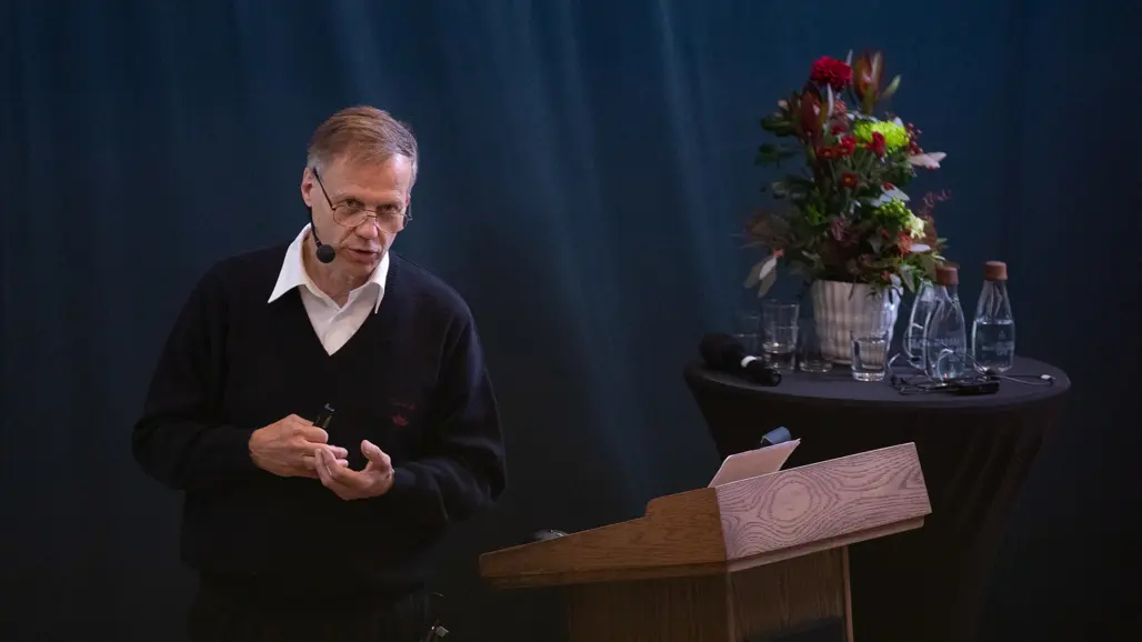 En man vid ett podium med blommor i bakgrunden. Foto.
