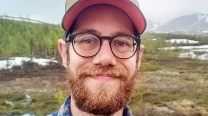 Bilden visar en man med skägg och glasögon som ler mot kameran. Han bär en keps med rosa insida och en rutig skjorta. Bakgrunden består av en naturskön miljö med gräs, snöfläckar och berg i fjärran, vilket tyder på en kall eller högfjällsmiljö.