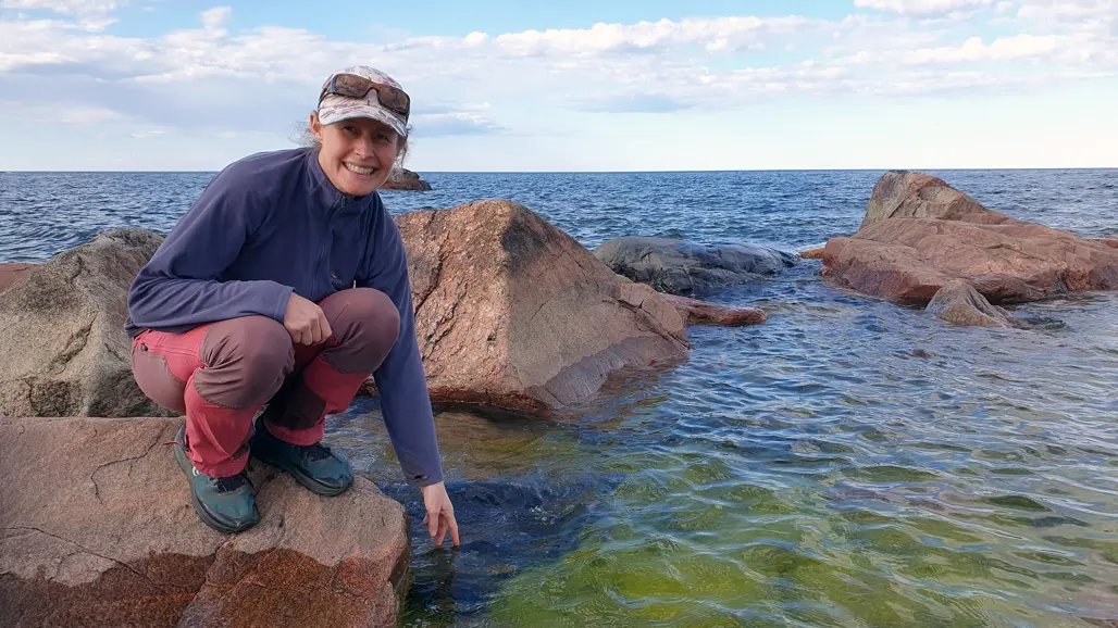 A smiling woman on a rock reaching her hand towards the water. Photo.