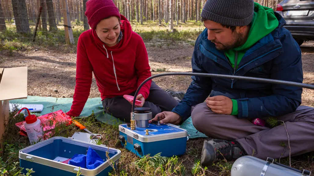 Två personer sitter på marken i skogen och arbetar med ett instrument i en blå låda