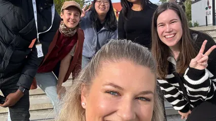 En grupp glada människor tar en selfie utomhus på en trappa. Personerna bär olika kläder, inklusive jackor och mössor, och en person gör en fredstecken. Bakgrunden är suddig med byggnader synliga.