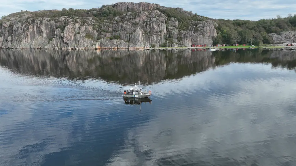 En vacker kustmiljö med en fiskebåt på lugnt vatten, omgiven av dramatiska klippformationer och en del små byggnader i bakgrunden.