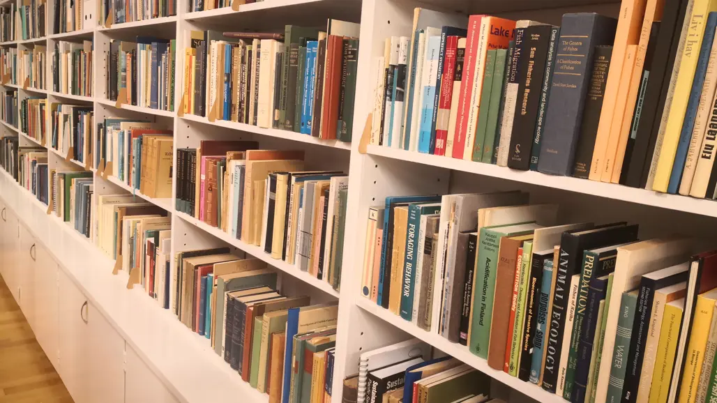 En lång vit bokhylla fylld med facklitteratur och vetenskapliga böcker, främst inom ekologi, vattenförvaltning och fiske. Böckerna står tätt packade i olika färger och storlekar. Golvet är av ljust trä, och hyllan har skåp med silverfärgade handtag längst ner.