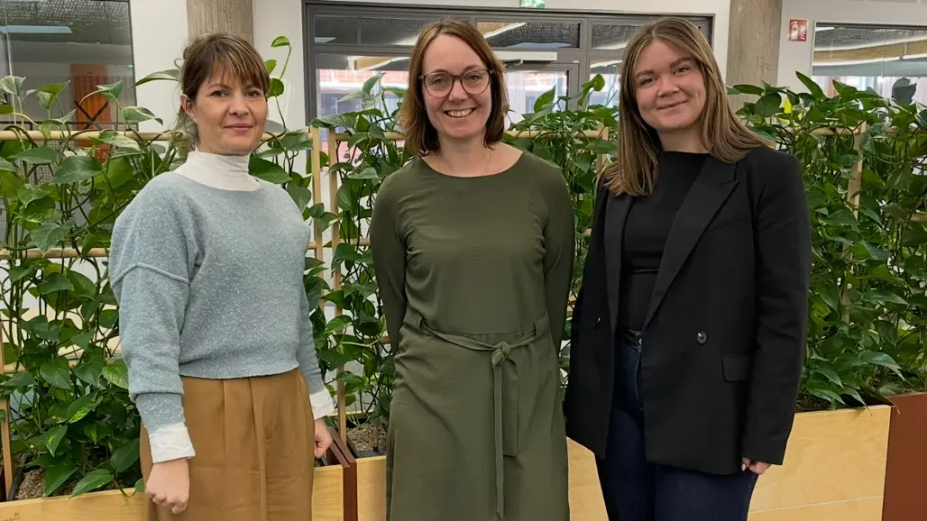 Tre kvinnor står tillsammans framför en rumsavskiljare med gröna växter
