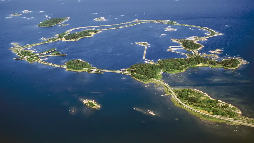 Flygbild över ett kustlandskap med flera sammanlänkade öar omgivna av blått hav. Smala vägar och broar förbinder öarna, som är täckta av grön vegetation och har små byggnader och bryggor. Mindre skär och holmar syns utspridda i vattnet runtomkring.