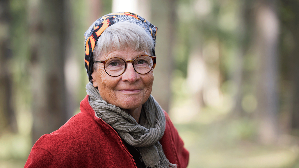 En äldre kvinna med halsduk, röd jacka och glasögon.