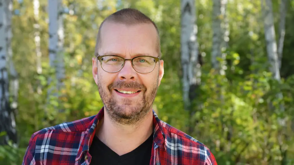 En man med glasögon och skäggstubb står med rödrutig skjorta i en björkskog.