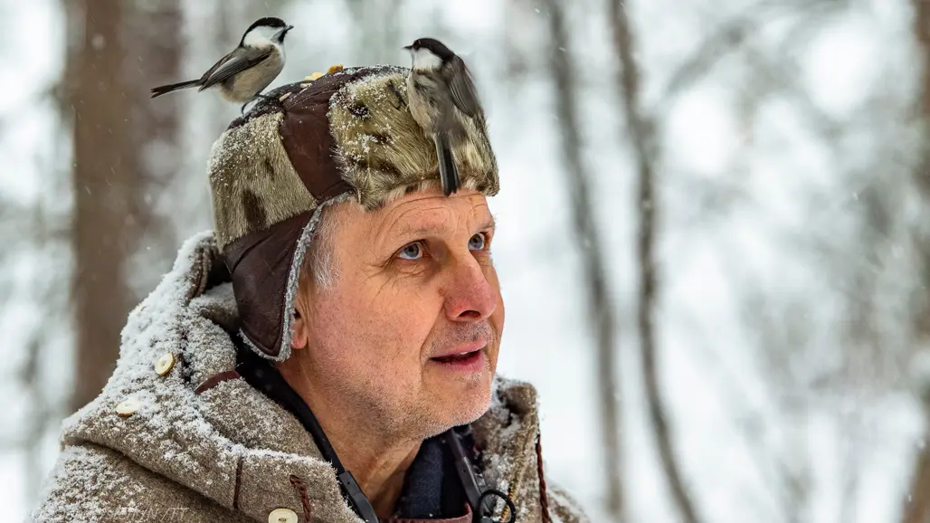 En person står i ett snöigt vinterlandskap och tittar uppåt. På huvudet har han en pälsmössa och på mössan sitter en liten fågel.
