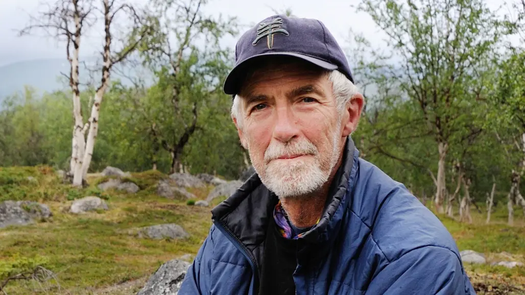 En friluftsklädd man med grått hår och skägg i ett fjällandskap, björkar med gula löv i bakgrunden.