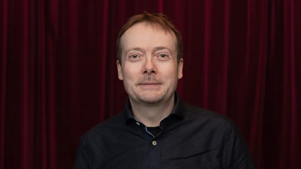 En person med kort hår och mörk skjorta sitter mot en vinröd bakgrund.