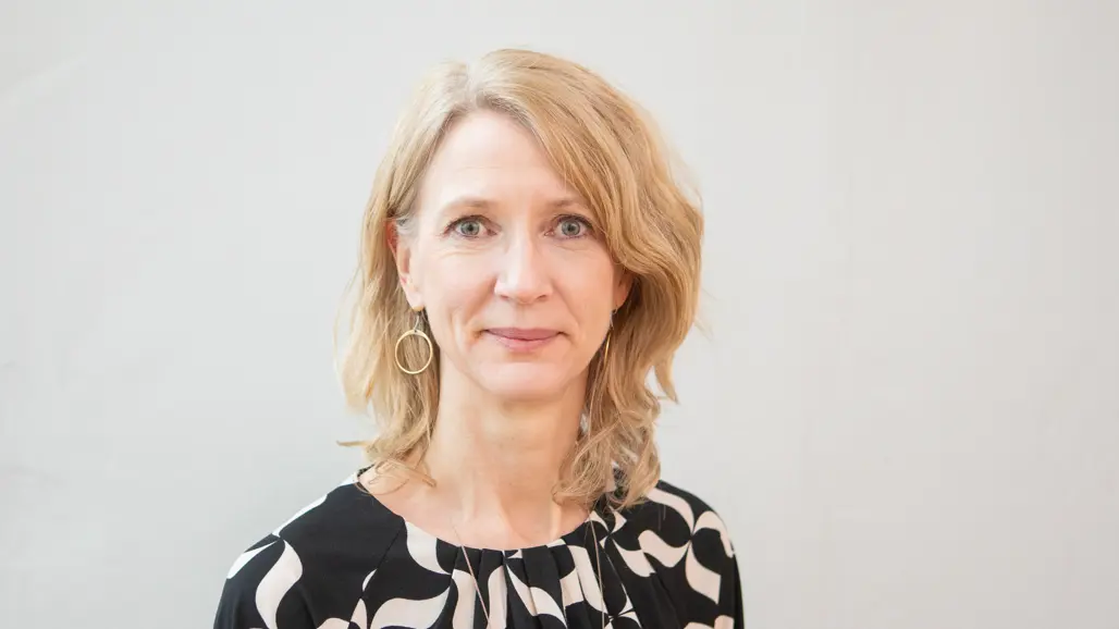 En leende blond kvinna med svart mönstrad blus.