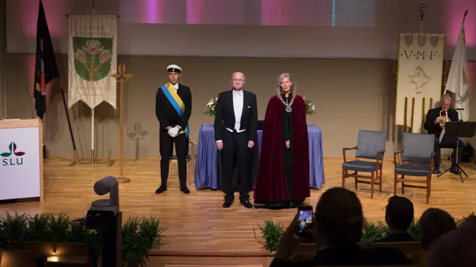 Två personer på en högtidlig scen, en man i frack och en kvinna med stor halskedja och röd kåpa i sammet.