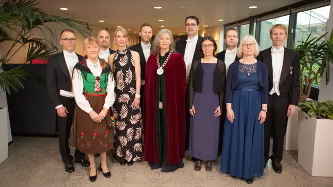 En gruppbild med elva högtidsklädda personer. En kvinna i rektorskedja och röd sammetskåpa står i mitten av gruppen.