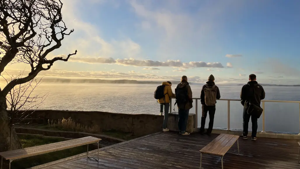 Fyra personer står vid ett räcke på en träveranda och blickar ut över ett hav eller en stor sjö. Solen är på väg ner eller upp, vilket skapar ett gyllene ljus på vattnet och himlen. En krokig trädgren syns till vänster i bilden, och två enkla bänkar står på verandan. Det är en kylig dag, då personerna bär varma jackor och ryggsäckar.