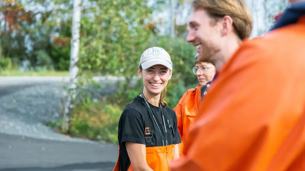 En leende person i keps och orange skyddsförkläde står utomhus och ser mot kameran. I bakgrunden syns fler personer i liknande klädsel, med träd och en grusväg i miljön bakom dem