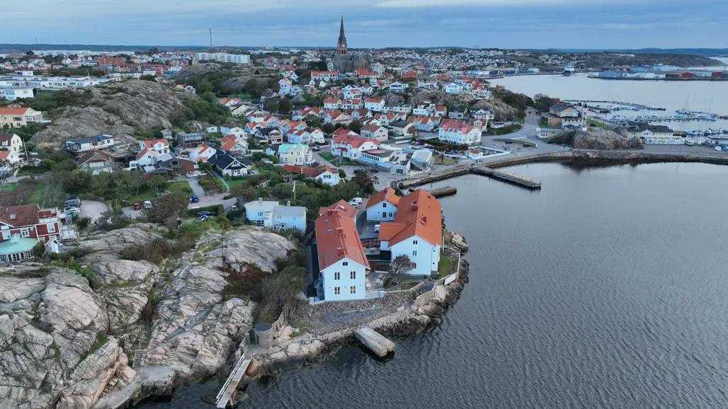 Flygbild över Havsfiskelaboratoriet i Lysekil med vita byggnader och rött tegeltak, belägen på en klippa vid vattnet. I bakgrunden syns en tätbebyggd skärgårdsstad med villor, en idrottshall och småbåtshamnar. Omgivningen består av karga klippor, grönska och hav.